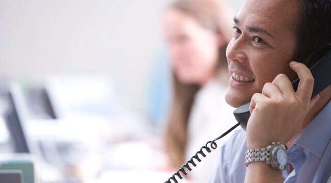 Muž v kancelárii drží telefón pri uchu, telefonuje a usmieva sa.